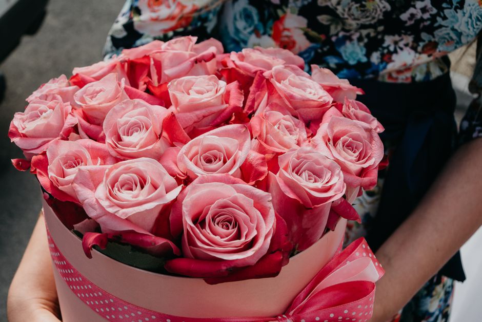 A round, light pink floral gift box filled with a dense arrangement of fresh, fully bloomed pink roses, each with delicate, velvety petals forming a spiral pattern. The roses are arranged tightly toge