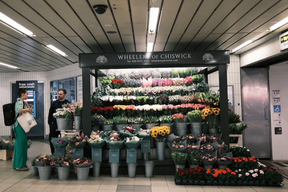 A vibrant flower stall named Wheeler of Chiswick located in a subway station, displaying a wide variety of fresh floral arrangements and bouquets. The flowers include bright yellow sunflowers, pink ro