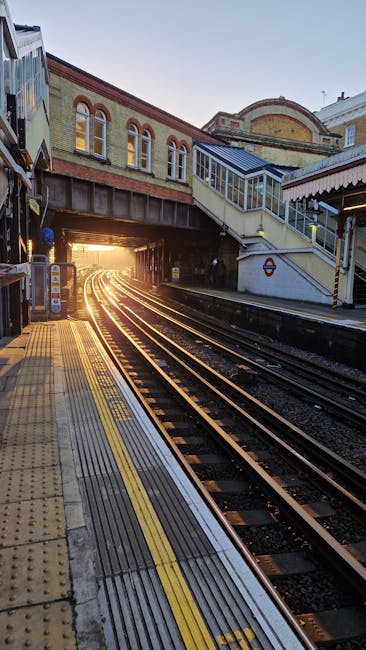The image depicts an outdoor railway station platform at sunset, with railway tracks curving into a tunnel illuminated by warm golden light. The platform features yellow tactile paving along the edge,