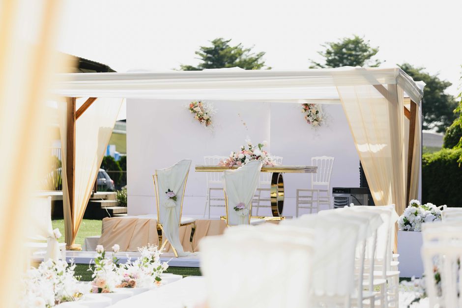 A floral arrangement featuring soft pink and white roses, complemented by delicate greenery, is placed on a rectangular table within a white outdoor wedding pavilion. The arrangement is surrounded by 