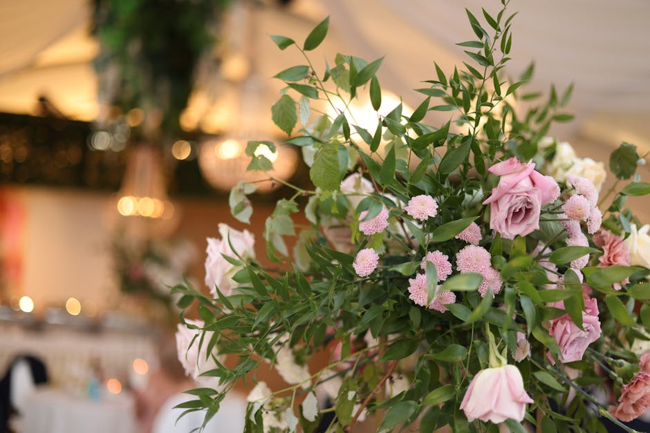 A close-up of a floral arrangement featuring soft pink roses, small pink daisy-like flowers, and lush green foliage with elongated leaves. The flowers are fresh, with delicate petals and firm stems, a
