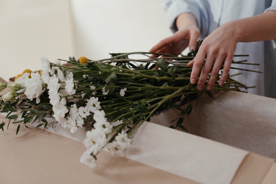 A person’s hands arranging a bouquet of fresh white carnations, yellow billy balls, and greenery with dark leaves onto a piece of beige wrapping paper. The flowers are vibrant and healthy, with deli
