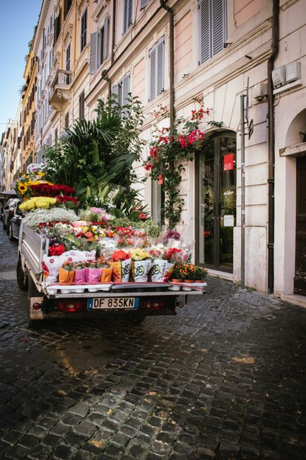 A small delivery truck parked on a cobblestone street in front of a building with cream-colored walls and large windows with shutters. The truck's bed is filled with a variety of fresh flowers, includ