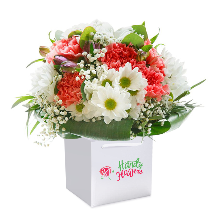 Arranged with exceptional care by knowledgeable Barkingside florists, this plush bouquet makes a bold statement as it stands tall within a refined white square gift bag, photographed against an immaculate white background to highlight every detail. Coral and peach carnations, plush and ruffled, form the focal point, their pastel tones gently transitioning into deep, vibrant reds-together embodying the lively character of Barkingside's bustling High Street. Amidst them, clusters of snowy white daisies with brilliant green-yellow centers impart a buoyant, sunny energy, reminiscent of the joyful scenery along the walking paths near Fairlop Waters. Gentle sprays of baby's breath add a misty softness throughout, balancing the palette with delicate, yet expressive notes. Tucked into the left side, a few purple-tipped closed buds add anticipation and visual depth. Framing the entire arrangement, broad, shaded green leaves provide a striking, glossy foundation, ensuring each flower stands out in perfect relief, much like the beloved green spaces that dot the Barkingside area. The bouquet's rounded, embracing silhouette is complemented by a sturdy white gift bag with soft ribbon handles, designed for comfort and ease of transport, particularly for swift delivery throughout Barkingside. Each floral element is arranged with both artistry and intent, creating a fresh, elegant statement piece ideal for celebrations, heartfelt thank-yous, or simply to spark delight at a special moment.