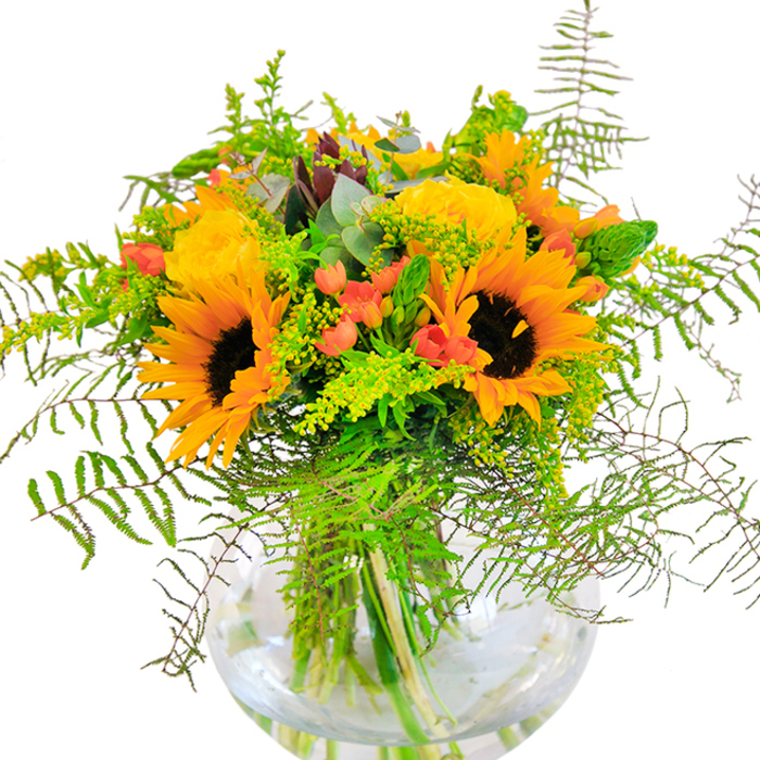 The first impression this exuberant arrangement gives is one of sunlight bottled into a bouquet for Barkingside homes. Large sunflowers shine at the center, their deep brown eyes smiling outward and petals glowing like rays over Fullwell Avenue. Intertwined amongst them, tightly petalled yellow garden roses unfurl in lush spirals, evoking both tradition and warmth. Pops of ruby red hypericum berries glisten among the leaves, while wispy ferns and stems of goldenrod spill gracefully from the boundaries, lending a feeling of movement and unstudied charm. Blue-green eucalyptus leaves appear at the edges, grounding the mix with cool, modern contrast. The spherical glass vase magnifies the cut stems, highlighting their arrangement and the refreshing clarity of the water-reminiscent of Barkingside's community displays. Every detail, from petal velvet to ferny softness, invites closer examination. The bouquet's faint fragrance and cheerful colours make it fitting for life's memorable moments-be it a joyful Barkingside birthday, a congratulatory gathering, or a simple act of appreciation. Crafted with care, it embodies the joy and vibrancy that define the local flower scene.