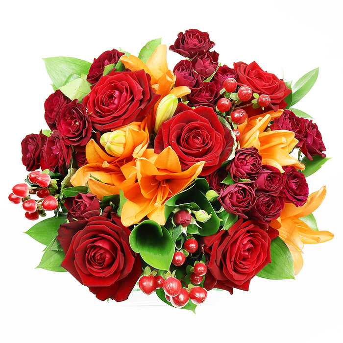From a top-down vantage against a spotless white surface, this bouquet presents an opulent, circular composition of deep, romantic reds and warm, sunlit oranges balanced by fresh greenery. Several substantial deep red roses form the visual nucleus, their velveteen petals unfurling into concentric folds that read as luxurious and tactile. Bright orange Asiatic lilies thread between these roses, their trumpet shapes creating dynamic lines; some lilies are opened wide, showing delicate speckling and erect stamens, while others remain in tighter bud forms, introducing a sense of unfolding time. Interspersed are tight clusters of dark burgundy spray roses, whose smaller heads add dense, velvety texture and a layered, jewel-toned shadow beneath the larger blooms. Lively red hypericum berries are tucked throughout, offering glossy spherical accents that bounce light and provide a festive, almost jewel-like punctuation. Lush green leaves-likely ruscus or salal-peek and fan around the edges, their clean glossy surfaces setting off the warm palette and lending the arrangement structural clarity. The soft, even lighting reveals subtle sheen and depth without harsh contrasts, inviting the viewer to imagine the bouquet's fragrance: rose's classic sweetness tempered by the lilies' spicy citrus note. Crafted with an eye for balance and seasonality, this bouquet suits anniversary celebrations, romantic gestures, or autumnal table displays, and would be equally at home delivered to a neighbour near Valentines Park or placed on a mantle in a Barkingside bungalow after a stroll through the local market.
