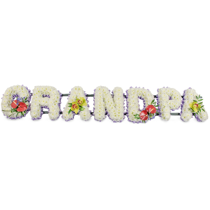 A long, elegant floral arrangement spells out GRANDPA in bold capital letters, resting against a plain white background that allows the craftsmanship and colours to stand out. Each letter is carefully constructed as a three-dimensional form, then completely covered with tightly packed white chrysanthemums, their small, delicate petals layered on top of one another to create a dense, padded effect. The white blooms give a sense of calm and serenity, their even texture suggesting a comforting softness despite the solid structure beneath. Just visible along the base of every letter is a neat trim of light lavender ribbon, providing a gentle thread of colour that outlines the shapes and adds a subtle decorative touch. A dark green bar runs behind the letters, linking them together and holding the word in a straight line, as you might see arranged along the front of a hearse or near the lectern at a Barkingside funeral service. Four accent sprays lend depth and individuality to the tribute. The 'G' on the left and the central 'D' feature clusters of rich red anthuriums, their glossy, heart-shaped spathes and golden spadices rising from a nest of dark green foliage, with tiny flecks of purple filler blooms adding extra interest. On the first 'A' and the final 'A', delicate yellow-green orchids are arranged with care, their graceful, arching petals and fresh, lime-toned colouring symbolising affection and remembrance. The overall design is dignified and expressive, a bespoke Grandpa floral tribute that families in Barkingside might send to honour a cherished grandfather, sharing love and respect through beautiful, thoughtfully chosen flowers.